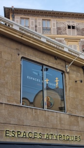 A building exterior featuring a large window reflecting an image of a religious icon and crosses. The structure has beige stone walls and detailed architectural elements with artwork and shutters seen on the upper level. The text 'ESPACES ATYPIQUES' is prominently displayed on the facade.