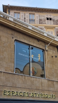 A building exterior featuring a large window reflecting an image of a religious icon and crosses. The structure has beige stone walls and detailed architectural elements with artwork and shutters seen on the upper level. The text 'ESPACES ATYPIQUES' is prominently displayed on the facade.