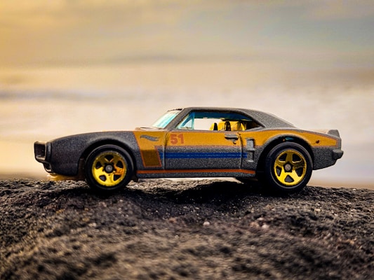 A toy car with a vintage design sits on a rough, textured surface against a backdrop of a soft, clouded sky. The car features a sleek body with a number '51' on the side, complemented by shiny gold-colored wheels, combining hues of black, gold, and orange.