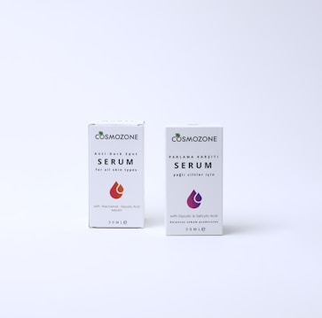 Two boxes of Cosmozone serum are displayed against a plain background. Each box features distinctive graphics indicating their specific purposes: one is an anti-dark spot serum for all skin types with niacinamide, glycolic acid, and arbutin, while the other targets oily skin with glycolic and salicylic acid for balancing sebum production.