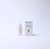 A bottle of Cosmozone serum with a dropper cap is placed next to its packaging box. The box features a golden droplet design, a green leaf icon, and text indicating anti-aging properties and ingredients such as hyaluronic acid, collagen, and niacinamide. Both items are on a plain, light background.
