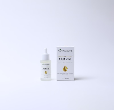 A bottle of Cosmozone serum with a dropper cap is placed next to its packaging box. The box features a golden droplet design, a green leaf icon, and text indicating anti-aging properties and ingredients such as hyaluronic acid, collagen, and niacinamide. Both items are on a plain, light background.