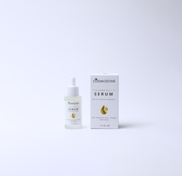 A bottle of Cosmozone serum with a dropper cap is placed next to its packaging box. The box features a golden droplet design, a green leaf icon, and text indicating anti-aging properties and ingredients such as hyaluronic acid, collagen, and niacinamide. Both items are on a plain, light background.