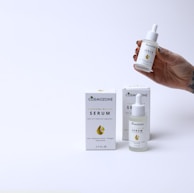 A hand holding a small dropper bottle labeled 'Cosmozone Serum' with two similar bottles and boxes displayed on a light background. The packaging contains text indicating the serum contains hyaluronic acid, collagen, and niacinamide.