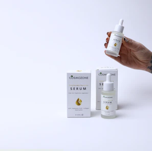 A hand holding a small dropper bottle labeled 'Cosmozone Serum' with two similar bottles and boxes displayed on a light background. The packaging contains text indicating the serum contains hyaluronic acid, collagen, and niacinamide.
