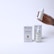 A hand holding a small dropper bottle labeled 'Cosmozone Serum' with two similar bottles and boxes displayed on a light background. The packaging contains text indicating the serum contains hyaluronic acid, collagen, and niacinamide.