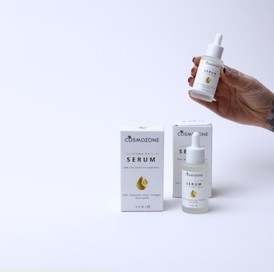 A hand holding a small dropper bottle labeled 'Cosmozone Serum' with two similar bottles and boxes displayed on a light background. The packaging contains text indicating the serum contains hyaluronic acid, collagen, and niacinamide.