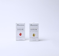 Two boxes of Cosmozone serum are placed on a light background. The box on the left is labeled 'Anti-Dark Spot Serum for all skin types' and features a red droplet graphic. The box on the right is labeled with non-English text for 'Serum' and includes a gold droplet graphic. Both boxes display additional product information and measure 30ML.