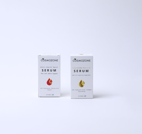 Two boxes of Cosmozone serum are placed on a light background. The box on the left is labeled 'Anti-Dark Spot Serum for all skin types' and features a red droplet graphic. The box on the right is labeled with non-English text for 'Serum' and includes a gold droplet graphic. Both boxes display additional product information and measure 30ML.