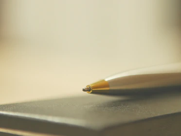 A close-up of a sleek metal ballpoint pen resting on a wooden desk with soft natural light.