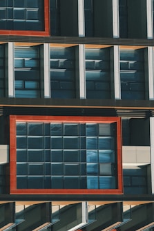 The image captures a section of a modern building facade featuring a grid of large, reflective windows. The window frames are accented with bold, red outlines that contrast with the darker surfaces of the building. Vertical and horizontal elements create a geometric pattern, and the overall architectural design emphasizes symmetry and modernity.
