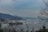 A large cargo ship is navigating through a wide body of water surrounded by hills. On one side, residential buildings and vegetation can be seen. The scene is overcast with bare tree branches framing the image.