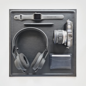 A neatly arranged flat lay featuring a smartwatch with a gray wristband, a stylus, a compact camera with a lens, a pair of black headphones, and a black wallet. These items are placed on a rectangular black leather mat.