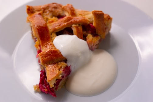 A slice of pie with a golden brown lattice crust sits on a white plate. The pie filling appears to be a vibrant red, possibly berry-flavored. Beside the pie, there is a dollop of white cream or yogurt.