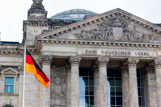 A grand architectural building with ornate carvings above the entrance and large stone columns supporting the structure. The phrase 'Dem Deutschen Volke' is engraved above the entrance. A German flag with black, red, and yellow stripes is prominently displayed in the foreground.