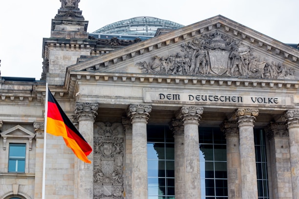 A grand architectural building with ornate carvings above the entrance and large stone columns supporting the structure. The phrase 'Dem Deutschen Volke' is engraved above the entrance. A German flag with black, red, and yellow stripes is prominently displayed in the foreground.