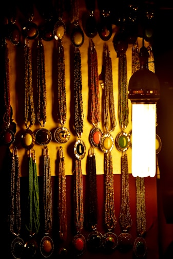 A dimly lit display of ornate necklaces with oval pendants featuring various colored stones is arranged on a wall. The necklaces are hanging in neat rows. A single, large hanging bulb provides warm illumination to the scene, casting soft shadows and accentuating the rich textures and colors of the jewelry.