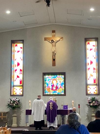 Inside a church, a crucifix hangs on the wall above a stained glass window depicting a religious figure. Two clergy members stand at an altar dressed in liturgical garments. Flanking them are stained glass windows with geometric designs and large floral arrangements. The seated congregation is partially visible in the lower foreground.