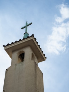 A tall, white bell tower is topped with a green cross. The tower has textured walls and an arched opening, with a small roof covered in tiles. The sky is partially cloudy with a light blue hue.