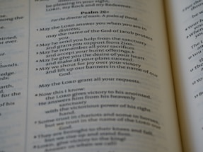 An open book displaying parts of Psalm 20 in clear and readable text. The pages are well-lit with a gentle focus, showcasing a passage with numbered verses. The text is printed in a serif font and is centered within the pages, revealing a religious context.