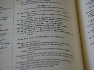 An open book displaying parts of Psalm 20 in clear and readable text. The pages are well-lit with a gentle focus, showcasing a passage with numbered verses. The text is printed in a serif font and is centered within the pages, revealing a religious context.