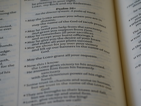 An open book displaying parts of Psalm 20 in clear and readable text. The pages are well-lit with a gentle focus, showcasing a passage with numbered verses. The text is printed in a serif font and is centered within the pages, revealing a religious context.