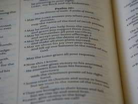 An open book displaying parts of Psalm 20 in clear and readable text. The pages are well-lit with a gentle focus, showcasing a passage with numbered verses. The text is printed in a serif font and is centered within the pages, revealing a religious context.