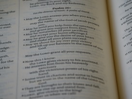 An open book displaying parts of Psalm 20 in clear and readable text. The pages are well-lit with a gentle focus, showcasing a passage with numbered verses. The text is printed in a serif font and is centered within the pages, revealing a religious context.