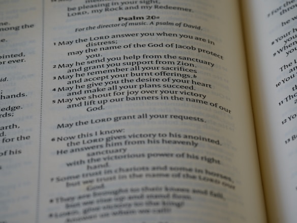 An open book displaying parts of Psalm 20 in clear and readable text. The pages are well-lit with a gentle focus, showcasing a passage with numbered verses. The text is printed in a serif font and is centered within the pages, revealing a religious context.