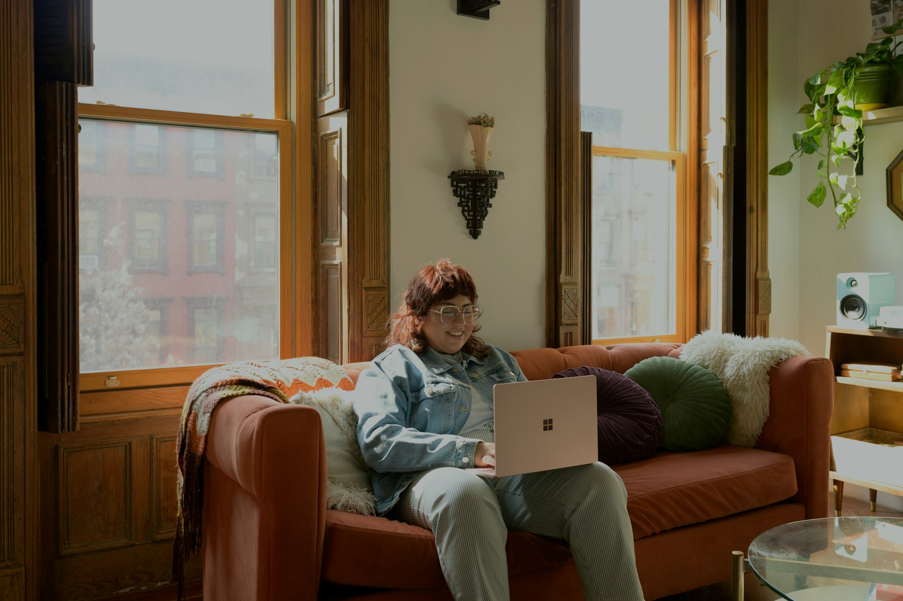 A woman sitting on a couch working on a laptop with a window behind her