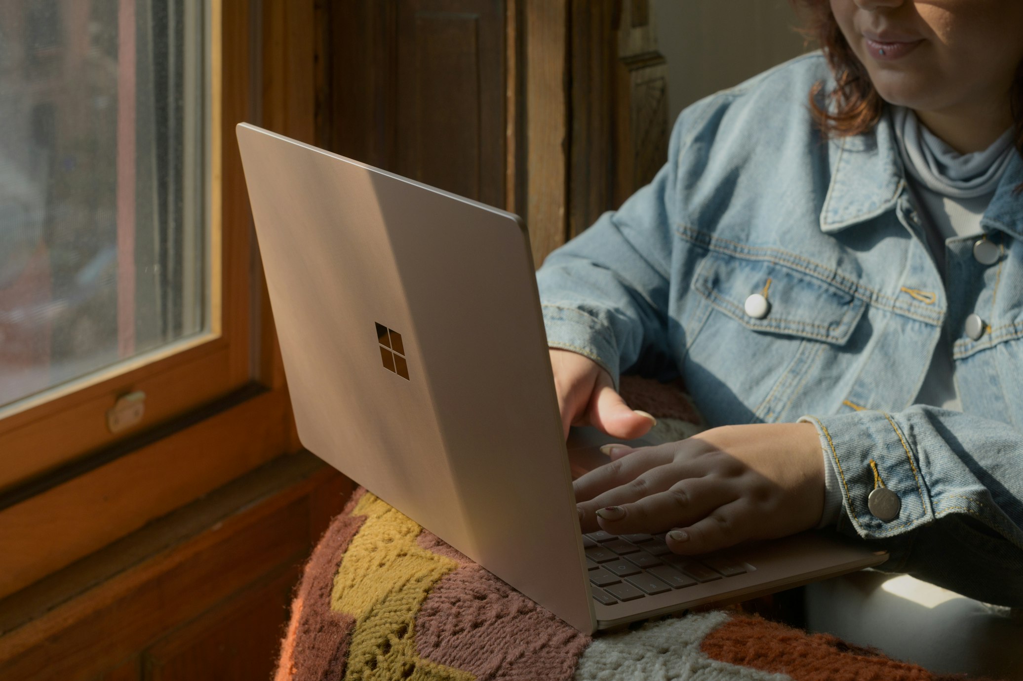 Person typing on rose gold laptop in warm sunlight near window