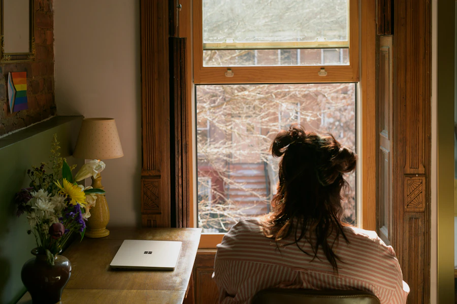 An adult sits by a sunlit window at a wooden desk seen from behind, warm afternoon light filling a calm room