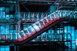 A futuristic escalator enclosed in a transparent tube runs diagonally across an industrial architectural setting. The scene is illuminated with vibrant neon blue and pink lights, casting reflections on the glass surfaces.