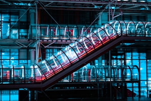 A futuristic escalator enclosed in a transparent tube runs diagonally across an industrial architectural setting. The scene is illuminated with vibrant neon blue and pink lights, casting reflections on the glass surfaces.