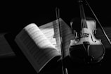 A violin and a bow are displayed alongside an open sheet music book, which is partially illuminated by a soft light. The scene is portrayed in grayscale, enhancing the shadows and contours of the instruments and papers.