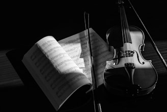 A violin and a bow are displayed alongside an open sheet music book, which is partially illuminated by a soft light. The scene is portrayed in grayscale, enhancing the shadows and contours of the instruments and papers.