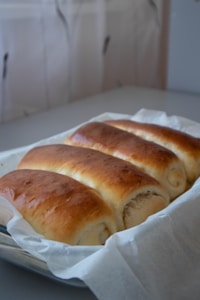 Four golden brown, freshly baked bread rolls rest in a baking dish lined with parchment paper. The setting appears to be a kitchen with soft lighting.