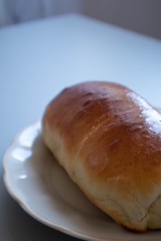 A freshly baked loaf of bread rests on a white ceramic plate placed on a light-colored table. The bread has a golden-brown crust with a glossy finish, indicating it has been baked to perfection. The setting appears simple and minimalist, focusing attention on the bread.