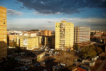 An urban landscape with multiple residential buildings, including a yellow high-rise and a distinctive hotel with a patterned facade. The scene is bathed in warm, golden sunlight, creating long shadows. Numerous parked cars are visible in the parking areas below, and there are trees scattered around. The sky is partly cloudy with a mix of blue and dark clouds.