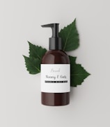 A brown bottle with a black pump dispenser is placed against a neutral, light background. The label on the bottle reads 'Brand Honey & Oats, Organic Body Wash' in a stylish font. Behind the bottle are several green leaves, adding a natural and organic touch to the composition.