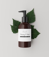 A brown bottle with a black pump dispenser is placed against a neutral, light background. The label on the bottle reads 'Brand Honey & Oats, Organic Body Wash' in a stylish font. Behind the bottle are several green leaves, adding a natural and organic touch to the composition.