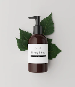 Softly lit close-up of a baby shampoo and body wash bottle surrounded by gentle green leaves.