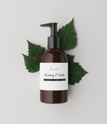 A brown bottle with a black pump dispenser is placed against a neutral, light background. The label on the bottle reads 'Brand Honey & Oats, Organic Body Wash' in a stylish font. Behind the bottle are several green leaves, adding a natural and organic touch to the composition.