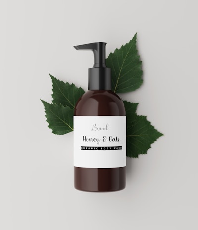 A brown bottle with a black pump dispenser is placed against a neutral, light background. The label on the bottle reads 'Brand Honey & Oats, Organic Body Wash' in a stylish font. Behind the bottle are several green leaves, adding a natural and organic touch to the composition.
