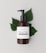 A brown bottle with a black pump dispenser is placed against a neutral, light background. The label on the bottle reads 'Brand Honey & Oats, Organic Body Wash' in a stylish font. Behind the bottle are several green leaves, adding a natural and organic touch to the composition.