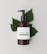 A brown bottle with a black pump dispenser is placed against a neutral, light background. The label on the bottle reads 'Brand Honey & Oats, Organic Body Wash' in a stylish font. Behind the bottle are several green leaves, adding a natural and organic touch to the composition.