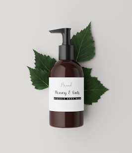 A brown bottle with a black pump dispenser is placed against a neutral, light background. The label on the bottle reads 'Brand Honey & Oats, Organic Body Wash' in a stylish font. Behind the bottle are several green leaves, adding a natural and organic touch to the composition.