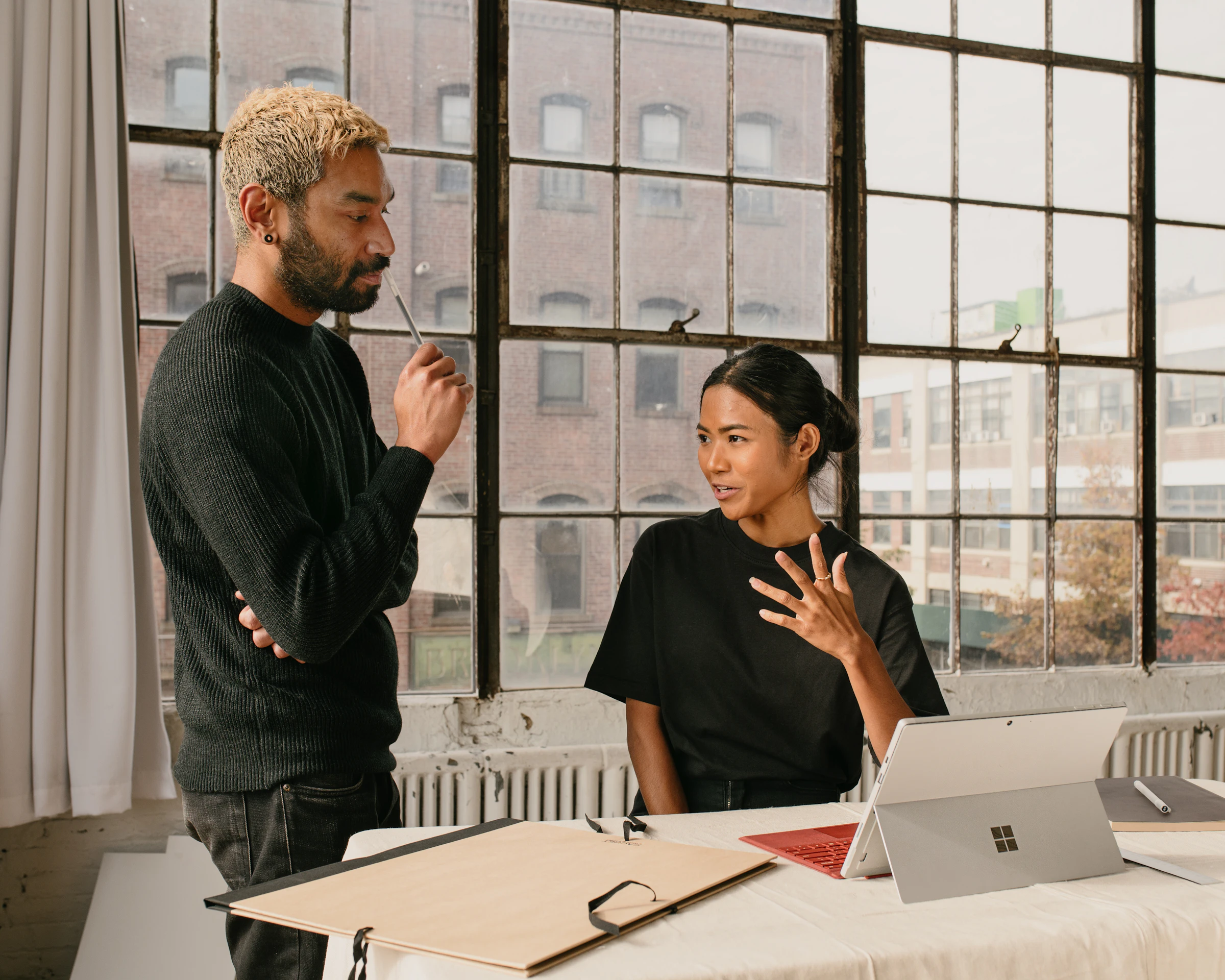 A woman explaining something with her hand raised while a colleague listens, beside tall loft windows looking onto brick buildings