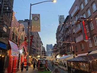 A narrow street lined with brick buildings is adorned with festive string lights and decorations. People walk along the street, which features restaurant signage and outdoor dining areas. Banners hang from the lamp posts, and the sky is transitioning to evening.