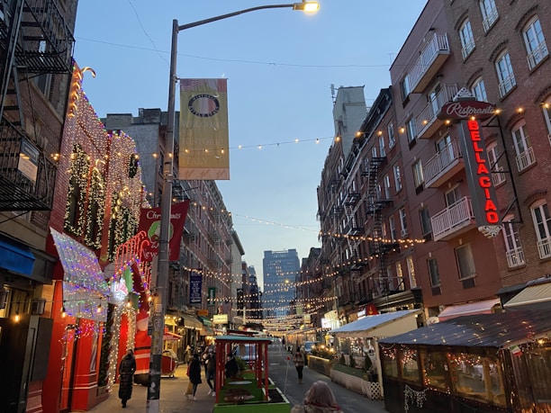 A narrow street lined with brick buildings is adorned with festive string lights and decorations. People walk along the street, which features restaurant signage and outdoor dining areas. Banners hang from the lamp posts, and the sky is transitioning to evening.